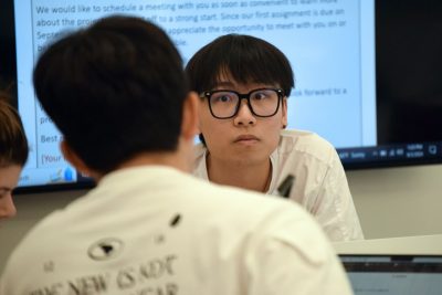 One student faces camera, while another has back to the camera. A projector screen is in the background.