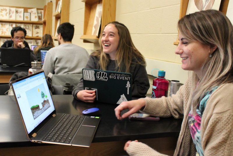 Two people sit in front of laptops, in a classroom of students.