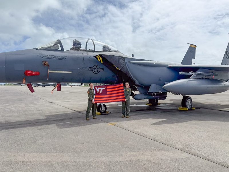 Stefaniak and Archer hold a striped VT flag. They are wearing military flight suits while smiling at the camera. A military fighter jet sits directly behind them. 