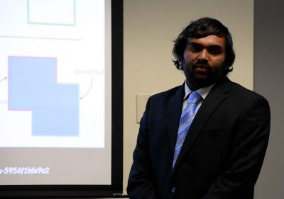 Student in suit stands in front of projection screen displaying a slide of capstone presentation.