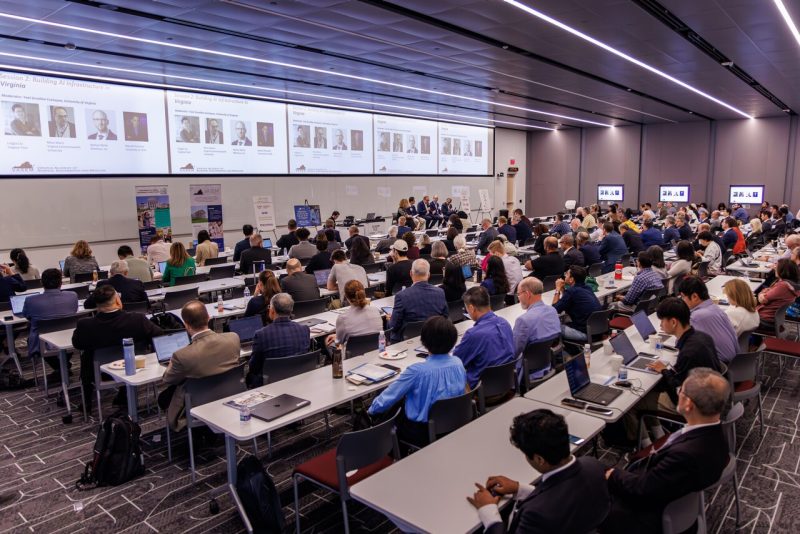 Room full of individuals at lined up tables with large screen displays at front of the room.