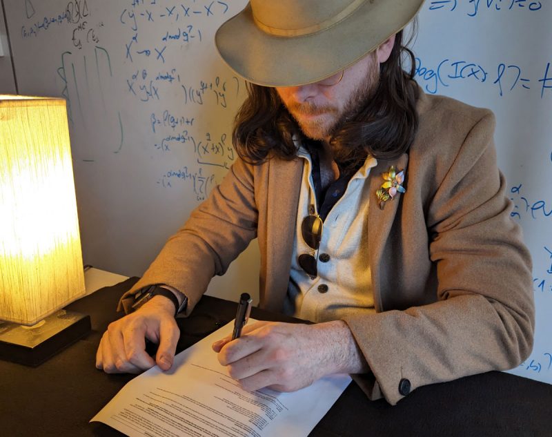 A man with a hat pulled low over his eyes signs a paper at a table. The whiteboard in the background is covered in equations.